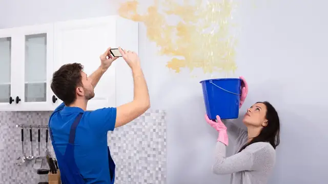 Homeowners photographing a water-stained wall while a bucket catches drips